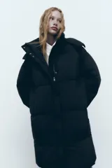 Cazadora larga acolchada negra con cuello alto y capucha desmontable con cremallera. Tiene manga larga con puño elástico interior, bolsillos delanteros con forro polar y cierre con botón a presión. Cierre frontal con cremallera oculta por solapa y botones a presión.