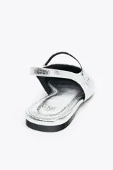 Zapato tipo bailarina destalonada plateada con efecto metalizado, punta redonda y tira delantera con aplique metálico.