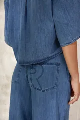 Camisa corta (crop) de denim azul, con calce oversize, cuello clásico, manga corta ancha y un bolsillo frontal.