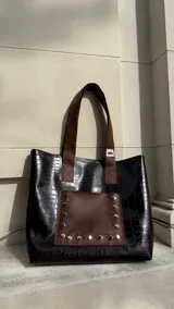 Bolso tote negro con textura símil cuero de cocodrilo, bolsillo marrón con tachas plateadas en el frente y asas de cuero marrón.