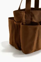 Bolso shopper marrón de lona de algodón con doble asa de mano y múltiples compartimentos exteriores, tres en los lados largos y uno de malla en los laterales.
