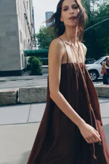 Vestido largo marrón de tejido satinado, con escote recto, tirantes finos extraíbles, bolsillos laterales ocultos y bajo asimétrico acabado en globo.