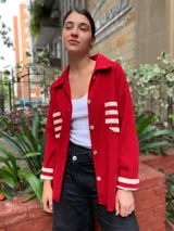 Camisa roja de tejido de algodón con cuello clásico, cierre frontal abotonado, mangas largas con puños acanalados a rayas y dos bolsillos de parche con rayas horizontales en el pecho.