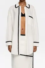 Camisa larga color blanco con borde de encaje negro, de corte recto, con cuello camisero, manga larga con puño y botón, y cierre frontal con botones.