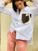 Camisa color naranja de lino o algodón con cuello clásico, cierre frontal con botones y mangas largas con puños abotonados. Presenta un bolsillo de cuero marrón en el pecho.