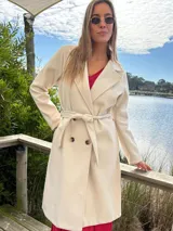Abrigo trench color crudo, de paño fino con dibujo en sarga, forrado, con cuello solapa, doble botonadura y cinturón con lazada.