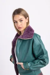 Chaqueta bomber de cuero verde esmeralda con cuello de piel sintética color lila, cierre frontal con cremallera, corte a la cintura, hombros caídos y mangas amplias.