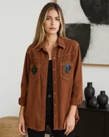 Camisa marrón de denim liviano con corte holgado, cuello clásico, cierre frontal con botones dorados, mangas largas con puños abotonados y ruedo desflecado. Presenta dos bolsillos de parche en el pecho con apliques de cuero negro en forma de rombo y tachas doradas.