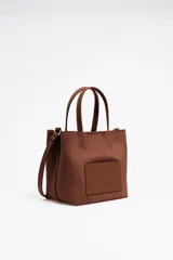 Bolso tote pequeño marca Bimba y Lola, color marrón, con doble asa de mano y asa bandolera de tejido. Presenta un bolsillo externo con el logo de la marca grabado.