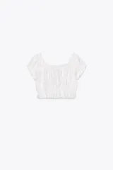Top crop blanco de escote cuadrado y manga corta abullonada. Detalle de lazada en el pecho y volantes en el escote y el bajo.