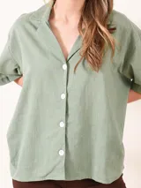 Camisa verde grisáceo de mangas cortas, cuello clásico y cierre frontal con botones blancos.