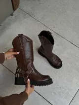 Bota de cuero vacuno engrasado color chocolate, con caña alta y textura desgastada. Presenta bordados hechos a mano, detalle de hebillas y agarre con avíos metálicos desmontables.