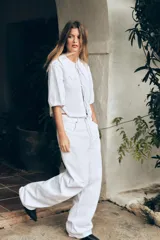 Camisa blanca de popelín con cuello bobo bordado y lazada. Tiene mangas abullonadas por debajo del codo con elástico y pinzas en la espalda. El cierre frontal es con lazadas.