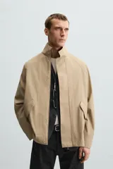 Parka ligera color beige, de corte relaxed fit, confeccionada en tejido técnico repelente al agua. Tiene cuello alto, mangas largas con puños ajustables con botón a presión, bolsillos de solapa en la cadera y cierre frontal con cremallera oculta por solapa con botones a presión.
