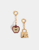Set de dos charms intercambiables dorados con mosquetón. Incluye un dije de cartera con moño y un dije de manzana esmaltada en rojo con strass.