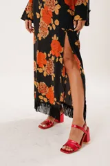 Vestido largo negro con estampado de flores naranjas, cuello mao con abertura, mangas largas acampanadas, espalda abierta, tajo frontal y flecos en el ruedo.