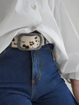 Cinturón con estampado de vaca en blanco y marrón, hebilla metálica rectangular con el nombre de la marca grabado.