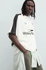 Camiseta blanca de corte holgado con mangas cortas negras. Presenta un logo azul en el pecho izquierdo, un escudo rojo con un caballo en el pecho derecho y la inscripción "Victory Games" en negro en el centro.