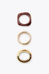 Pack de tres pulseras rígidas de diferentes tamaños, materiales y formas en relieve: una marrón cuadrada, una beige con forma irregular y una dorada con cierre.