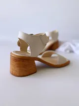 Sandalias de cuero color crudo, con tira ancha sobre los dedos, pulsera al tobillo con hebilla metálica y taco de madera de 6 cm de altura.