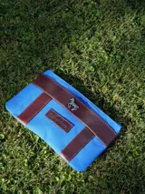 Sobre de lona azul con terminaciones en cuero marrón, cierre superior y aplique metálico de caballo en el frente.