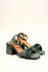 Sandalia de terciopelo verde con taco cuadrado de 7 cm, punta cuadrada y tiras anchas anudadas en el empeine. Cuenta con una tira fina ajustable al tobillo con hebilla dorada.