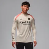 Remera de manga larga color crema con detalles en salmón en los hombros, con logo de Jordan y escudo del Paris Saint-Germain en el pecho y leyenda "PARIS" estampada verticalmente en la manga izquierda.