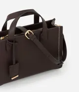 Cartera tipo satchel estructurada de color marrón oscuro, con costuras decorativas en contraste color crema en los laterales. Posee asas superiores cortas y una correa larga ajustable y desmontable. Los herrajes son de color dorado.