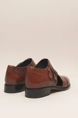 Zapato bajo de cuero negro, estilo monk strap con dos hebillas ajustables sobre el empeine. Presenta detalles de brogue (perforaciones) en la puntera y alrededor del borde superior. Tiene un taco bajo y ancho.