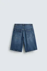 Bermuda de jean azul con lavado medio, corte holgado, cinco bolsillos, roturas y cierre frontal con cremallera y botón.
