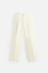 Pantalón color crema de lino, corte amplio, con pliegues frontales, bolsillos laterales y traseros de ojal. Cierre frontal con cremallera y gancho metálico oculto.