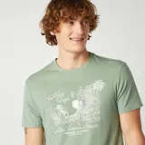 Camiseta verde de manga corta con cuello redondo y estampado blanco con la frase "Nice Trip" y un dibujo de edificios y palmeras.