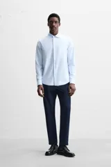 Camisa celeste a rayas verticales blancas, de corte recto, con cuello italiano abierto, manga larga con puños abotonados y cierre frontal con botones.