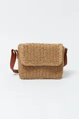 Cartera tipo bandolera de paja color beige con correa de cuero marrón ajustable.
