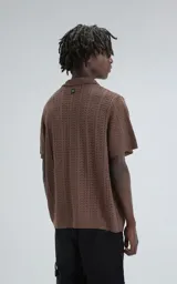 Camisa de tejido de punto color café, manga corta y cuello polo con botones frontales. Presenta un patrón calado de espiga o chevron.