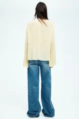 Blusa color crema de gasa con escote en V y cordones cruzados. Tiene mangas largas acampanadas y pliegues en la cintura.