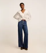 Blusa color blanco confeccionada en voile, de mangas largas con elástico en los puños, cuello en V con volado y lazo con adornos metalizados.