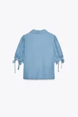 Camisa celeste de denim con cuello solapa, escote en pico, mangas abullonadas con lazos y cierre frontal con botones.