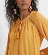 Blusa de manga larga color mostaza con textura de puntos en relieve (dobby). Presenta cuello redondo con abertura en V y cordón ajustable con borla (tassel) en el escote. Las mangas tienen detalles de entretejido calado (entremedios) y volados con terminación fruncida en los puños.