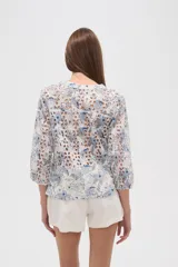 Blusa blanca de algodón con bordado broderie inglés y estampado floral en tonos azules y marrones. Posee cuello en V con abertura y botones frontales, y mangas tres cuartos.