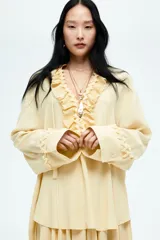 Túnica color amarillo pálido de corte holgado, con cuello mao y abertura frontal con volados plisados. Mangas largas con puños anchos con volados y botones. Bajo recto.