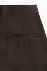 Pantalón ancho color marrón oscuro, de tiro alto con cierre, botón y corchetes ocultos, bolsillos al bies y bolsillos atrás decorativos. Piernas amplias con pliegue adelante y atrás.
