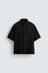 Camisa negra de manga corta con cuello solapa y corte relaxed fit. Presenta un patrón geométrico jacquard en toda la prenda, bajo con aberturas laterales y cierre frontal con botonadura oculta.