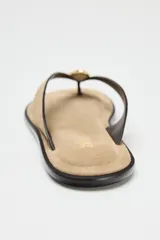 Sandalia plana color beige, con tira de serraje para el dedo y adorno metálico dorado.
