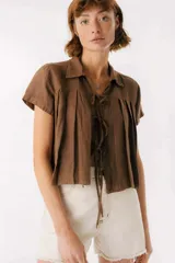 Camisa marrón de lino con cuello clásico, mangas cortas con dobladillo y cierre frontal con lazos. Presenta pinzas verticales en el frente.