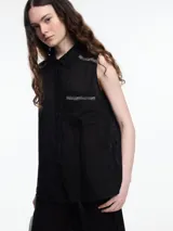 Camisa sin mangas negra de lino con cuello clásico, cierre frontal abotonado y bolsillo de parche en el pecho. Presenta bordados tipo pespunte en contraste en el frente y canesú trasero.