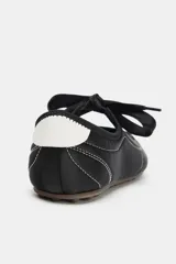 Zapato plano tipo bailarina de estilo deportivo, color negro, con pespuntes blancos contrastantes en el cuerpo y puntera. Presenta un escote redondo y un lazo decorativo en el empeine. La suela es fina y de color marrón claro.