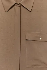 Camisa color marrón claro de cuello solapa y manga larga con trabilla y botón para remangar. Tiene un bolsillo de plastrón en el delantero, bajo asimétrico y cierre frontal con botones ocultos por solapa.