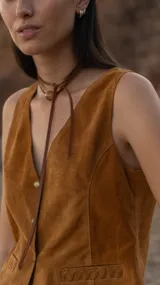 Chaleco marrón de gamuza con cuello en V, cierre con botones y bolsillos frontales con detalle de costuras. Corte recto.