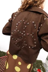 Chaqueta marrón de algodón con botones metálicos, cuello clásico, bolsillos frontales y apliques de tachas y botones formando una flor en la espalda.